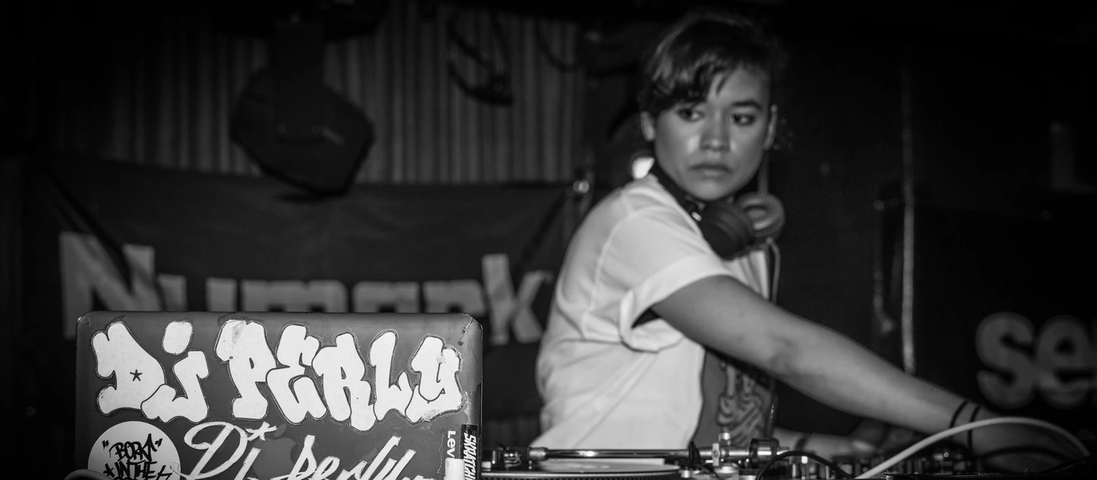 DJ Perly behind the turntables at a live set, her DJ Perly tag on the mixer flight case in frame