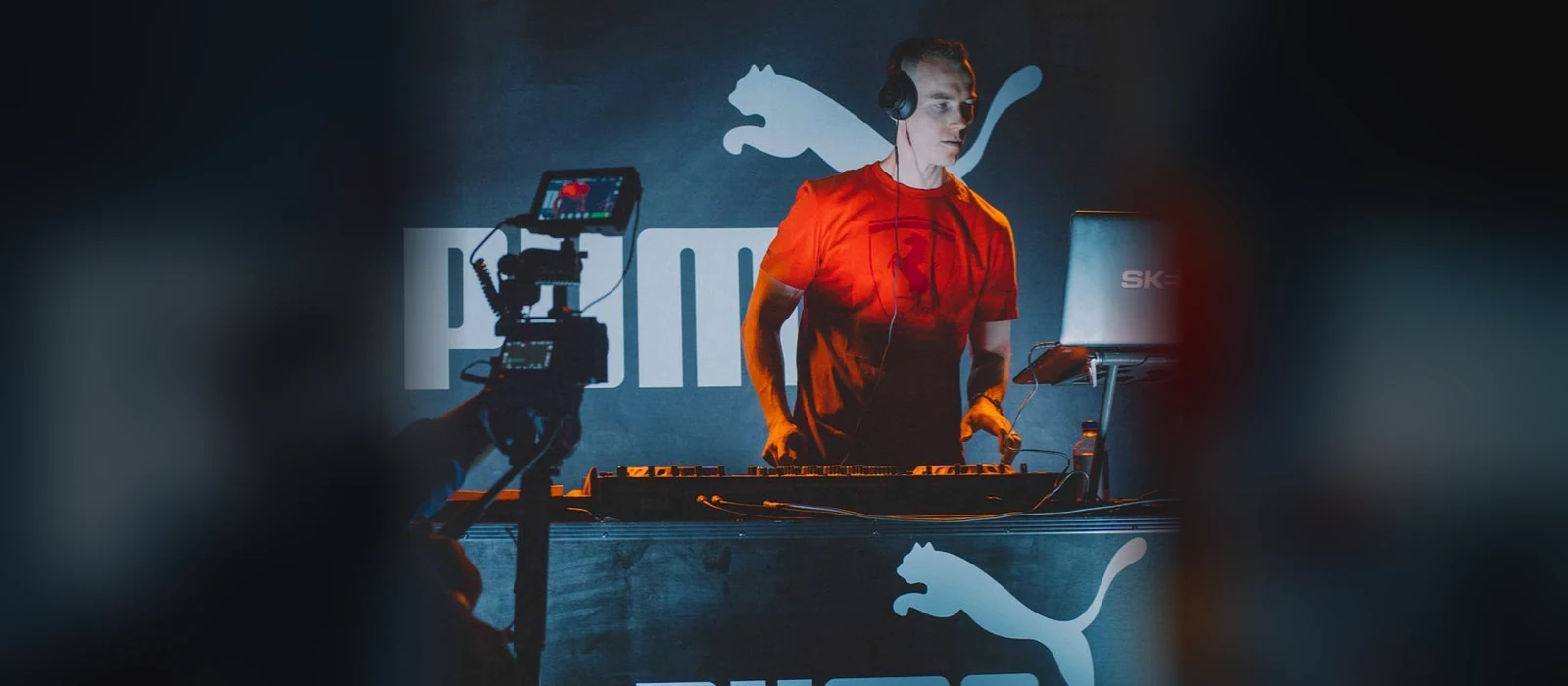 DJ Skee behind the decks at a PUMA event, orange tee, headphones on, the PUMA wordmark and cat logo on the wall behind him